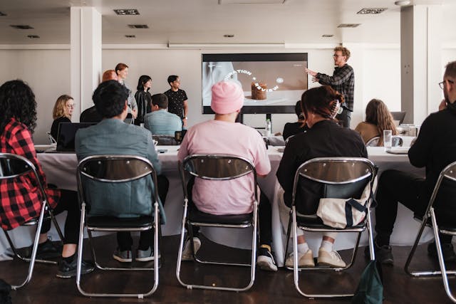 Man sieht einige Personen von hinten auf Stühlen sitzen. Sie hören einem Mann vor, der eine Powerpoint präsentiert